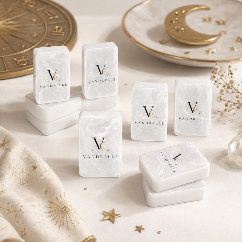 Puede incluir: Una colecci&oacute;n de bloques rectangulares con efecto de m&aacute;rmol blanco que presentan el texto negro VANDRELLE y un logotipo. Los bloques est&aacute;n dispuestos en pilas e individualmente sobre una superficie, acompa&ntilde;ados de una rueda zodiacal dorada y un plato de media luna.