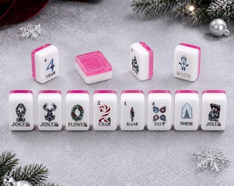 Könnte beinhalten: Ein Set aus weißen Mahjong-Spielsteinen mit pinkfarbenen Glitzer-Oberseiten, verziert mit weihnachtlichen Motiven und Schriftzügen wie Joker, Flower, Crak, Bam, Dot, North und White. Die Spielsteine sind in zwei Reihen auf einer grauen Fläche angeordnet.