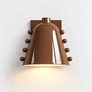 Pode incluir: Um candeeiro de parede castanho brilhante com um abajur em forma de sino e acabamento liso. A luminária é decorada com quatro pequenos botões esféricos dispostos verticalmente em cada lado do abajur e emite luz quente pela abertura inferior.