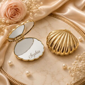 May include: Two gold-coloured metal compact mirrors shaped like seashells. One mirror is open, revealing a reflective surface with the name Sophia engraved in cursive script. The other mirror is closed, displaying a ribbed, scalloped shell exterior.