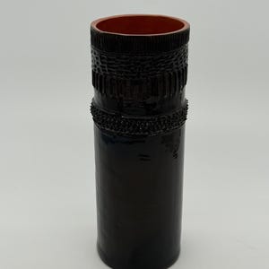 Op de afbeelding: Een hoge cilindrische keramische vaas met een glanzende zwarte buitenkant. Het bovenste gedeelte heeft een textuurband met verticale lijnen en een verhoogde, bobbelige ring. De binnenkant van de vaas heeft een contrasterende matte terracotta oranje kleur.