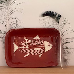 May include: A rectangular red ceramic tray featuring a white graphic illustration of a fish with patterned scales and fins. The fish design includes circular and wavy line details. Two long, thin, dark-tipped feathers are positioned behind the tray.