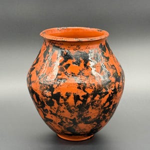 Pode incluir: Um vaso de cerâmica arredondado com acabamento brilhante, apresentando uma cor de base laranja vibrante e um padrão preto irregular e manchado que cobre a superfície externa. O vaso tem uma boca larga e aberta com uma borda ligeiramente alargada e uma base estreita.