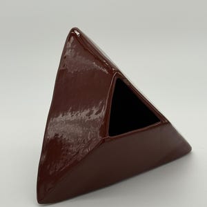 Könnte beinhalten: Eine glänzende, dunkel kastanienbraune Keramikvase in Form eines dreieckigen Prismas. Das Objekt hat eine glatte, reflektierende Oberfläche mit einer tief rotbraunen Glasur und eine hohle, dreieckige Öffnung an der Oberseite.