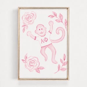 Op de afbeelding: Een ingelijste aquarelprint met een roze aap die een wit T-shirt draagt met de Griekse letters Alpha Phi in het roze gedrukt. De aap is omringd door roze aquarelrozen en bladerrijke takken op een witte achtergrond in een gouden lijst.