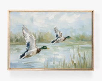 May include: An oil painting in a thin gold frame depicting two mallard ducks in flight over a marshy landscape. The scene features visible brushstrokes in shades of blue, green, and white, capturing the birds above calm water and tall reeds.