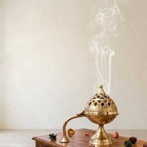 May include: A metallic gold-toned incense burner with a dome-shaped lid featuring intricate geometric cutouts. The burner has a long, curved handle and a pedestal base, with thin wisps of white smoke rising from the top of the structure.