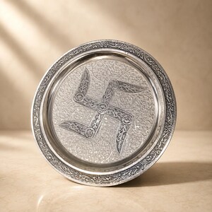 May include: A circular silver-toned metal tray featuring an intricate embossed swastika symbol at the center. The surface is decorated with detailed floral scrollwork patterns, and the raised rim has a repeating leaf-like border design.