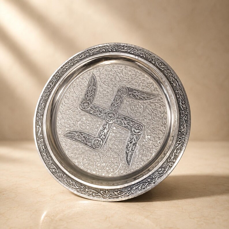 May include: A circular silver-toned metal tray featuring an intricate embossed swastika symbol at the center. The surface is decorated with detailed floral scrollwork patterns, and the raised rim has a repeating leaf-like border design.