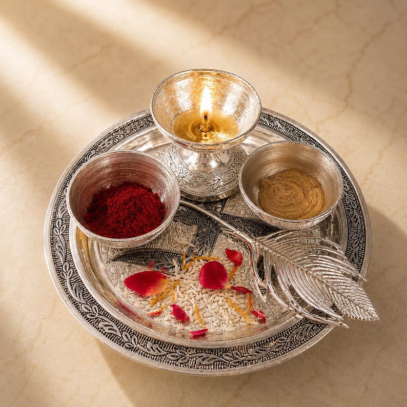 May include: A silver-toned metal puja thali set featuring an ornate circular tray, a lit diya lamp, two small bowls containing red powder and a tan paste, and a decorative feather-shaped spoon, all arranged with scattered white rice and rose petals.