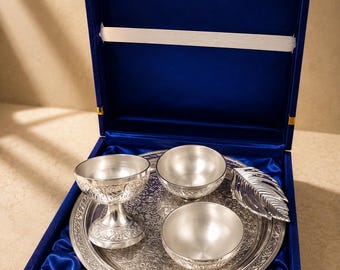 May include: A silver-toned metal set featuring a round, intricately engraved tray, two small bowls, one pedestal bowl, and a leaf-shaped spoon. The items are arranged inside a square, royal blue velvet-lined presentation box with a gold latch.