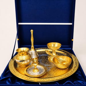 May include: A gold and silver-toned metal puja set arranged on a round tray inside a blue velvet-lined box. The set includes a small pot, a bell, a diya lamp, a bowl, a spoon, and a small plate, all featuring intricate floral engravings.