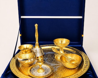May include: A gold and silver-toned metal puja set arranged on a round tray inside a blue velvet-lined box. The set includes a small pot, a bell, a diya lamp, a bowl, a spoon, and a small plate, all featuring intricate floral engravings.