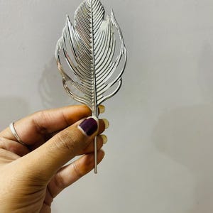 May include: A silver-colored metal hair accessory shaped like a stylized leaf or feather. The object features a central spine with symmetrical, cut-out vein patterns radiating outward, ending in a thin, straight pin at the bottom for securing hair.