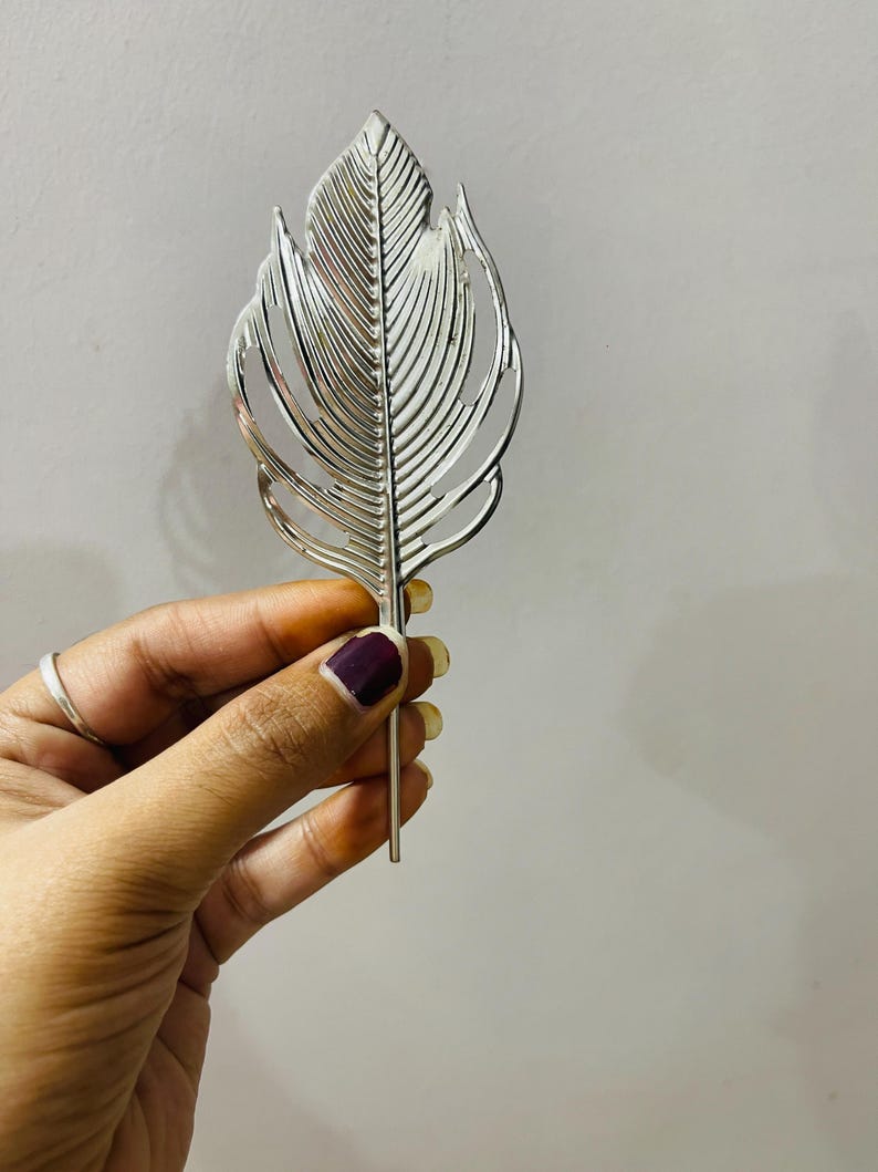 May include: A silver-colored metal hair accessory shaped like a stylized leaf or feather. The object features a central spine with symmetrical, cut-out vein patterns radiating outward, ending in a thin, straight pin at the bottom for securing hair.