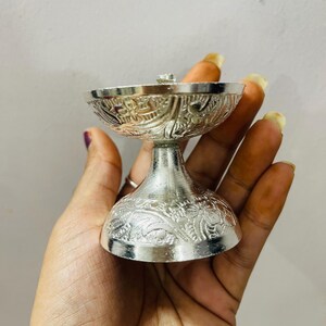 May include: A silver-colored metal oil lamp featuring an ornate, embossed floral pattern on both the wide, bowl-shaped top and the circular, flared base. The lamp has a smooth, polished finish and a distinct pedestal stem connecting the two sections.