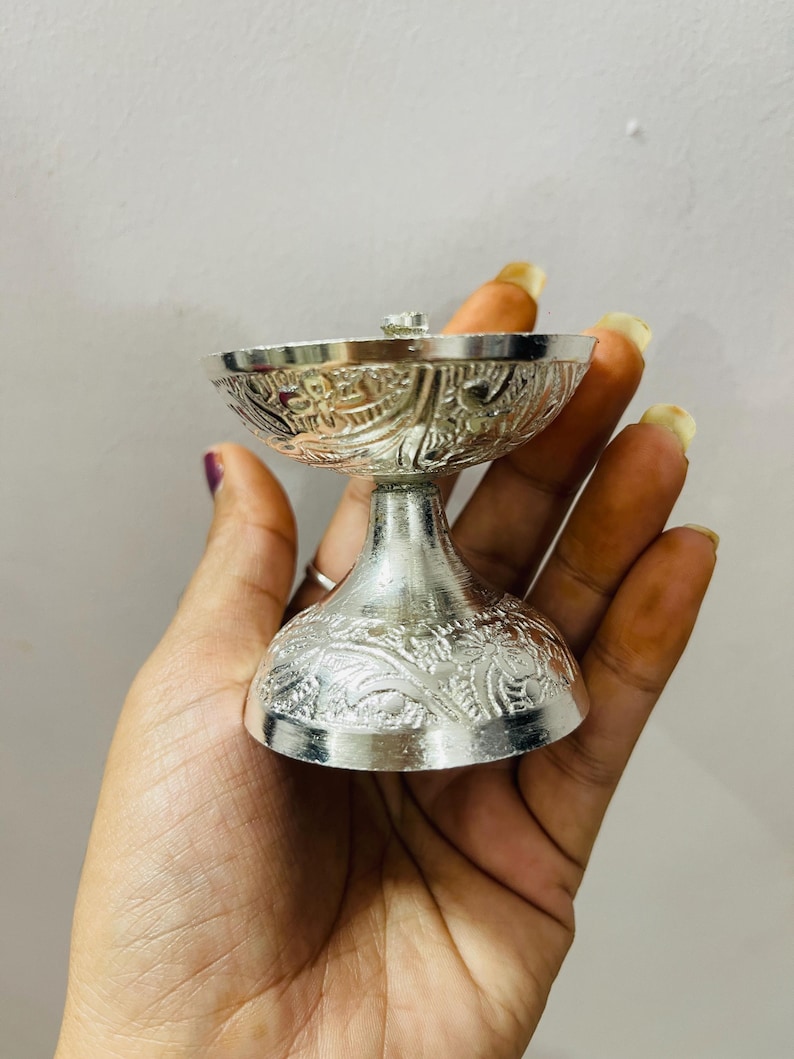 May include: A silver-colored metal oil lamp featuring an ornate, embossed floral pattern on both the wide, bowl-shaped top and the circular, flared base. The lamp has a smooth, polished finish and a distinct pedestal stem connecting the two sections.