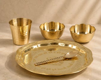May include: A polished brass dinnerware set consisting of a round plate with engraved text, a spoon, two scalloped bowls, and a cylindrical tumbler. The plate features a central inscription in Devanagari script and a decorative border pattern.