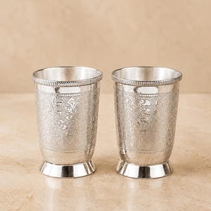 Könnte beinhalten: Zwei silberfarbene Metallbecher mit einem detaillierten, geprägten Blumenmuster auf der Außenfläche. Jeder Becher hat einen glatten, ausgestellten Boden und ein strukturiertes, seilartiges Randdetail am oberen Ende des zylindrischen Gefäßes.