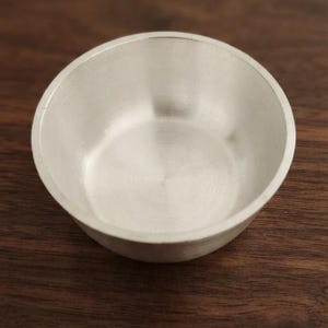 May include: A small, circular, silver-colored bowl with a smooth, metallic finish and a simple, open design. The bowl features a consistent, reflective surface with subtle circular brush marks visible along the interior base and walls.