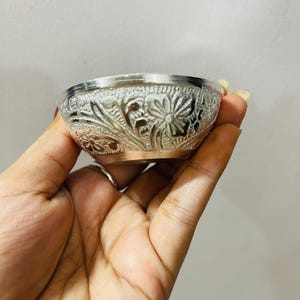 May include: A small, round, silver-colored bowl featuring an intricate, embossed floral pattern across its exterior surface. The bowl has a smooth, metallic rim and a visible copper-toned base, showcasing detailed craftsmanship and texture.