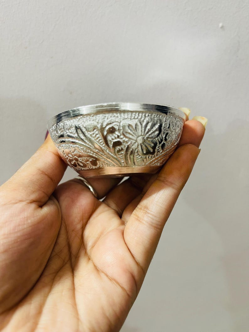 May include: A small, round, silver-colored bowl featuring an intricate, embossed floral pattern across its exterior surface. The bowl has a smooth, metallic rim and a visible copper-toned base, showcasing detailed craftsmanship and texture.