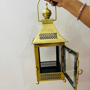 Puede incluir: Un farol de metal dorado con acabado envejecido, que presenta una puerta de cristal con bisagras, un remate decorativo en la parte superior y patrones recortados intrincados en las secciones superior e inferior. Incluye un asa resistente.
