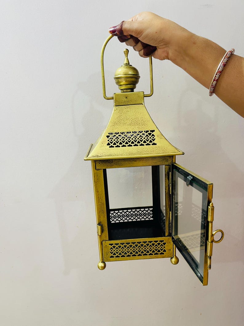 Puede incluir: Un farol de metal dorado con acabado envejecido, que presenta una puerta de cristal con bisagras, un remate decorativo en la parte superior y patrones recortados intrincados en las secciones superior e inferior. Incluye un asa resistente.