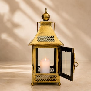 May include: A gold-coloured metal lantern with a distressed finish, featuring a pointed roof, a decorative lattice pattern, and a hinged door. Inside the lantern sits a single white pillar candle with a glowing flame, visible through the glass.