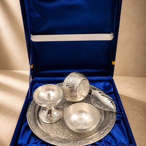 May include: A silver-colored metal set including a round tray with intricate floral engravings, a pedestal diya lamp, a small bowl, a larger embossed bowl, and a leaf-shaped spoon, all presented inside a blue velvet-lined display box.