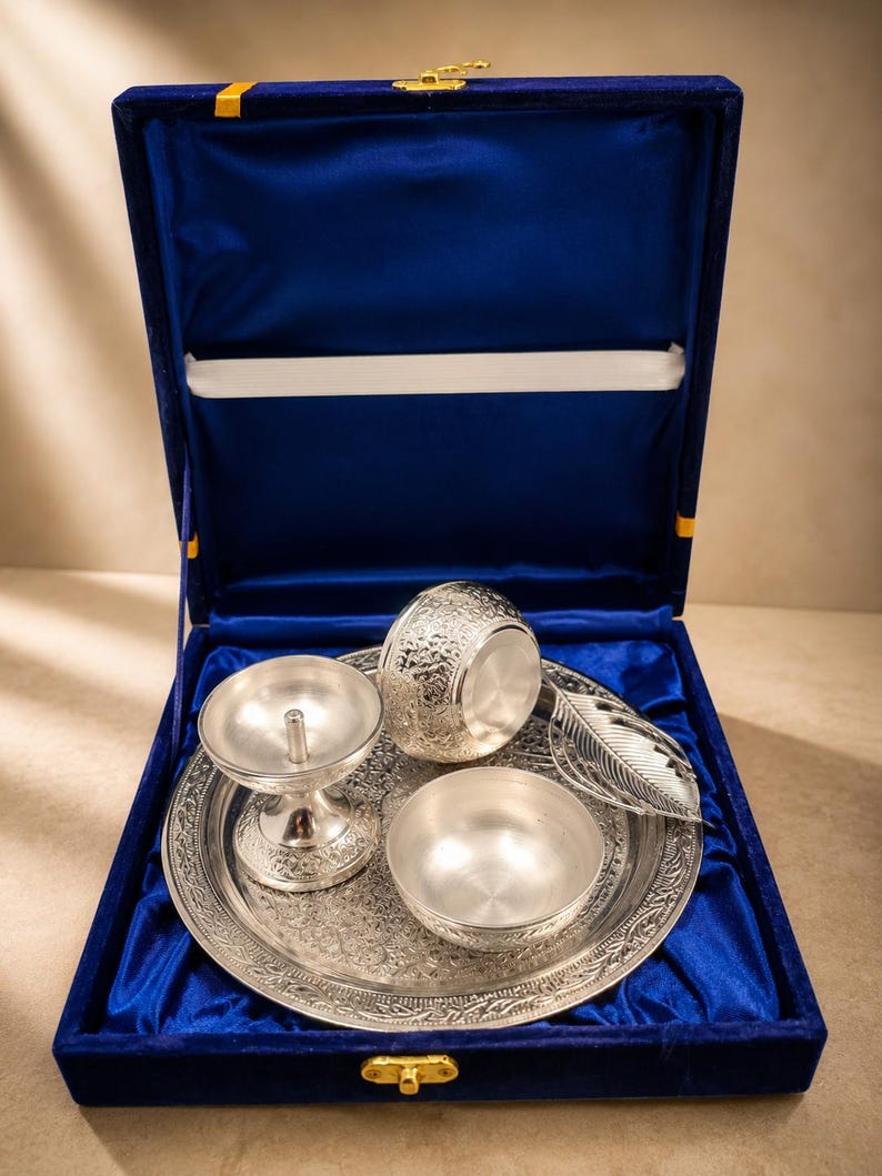 May include: A silver-colored metal set including a round tray with intricate floral engravings, a pedestal diya lamp, a small bowl, a larger embossed bowl, and a leaf-shaped spoon, all presented inside a blue velvet-lined display box.