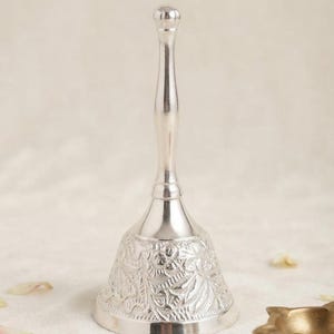May include: A silver-colored metal hand bell with a long, slender handle and a flared base. The bell features an intricate, embossed floral pattern covering the entire exterior surface of the flared section, ending in a smooth, rounded top knob.