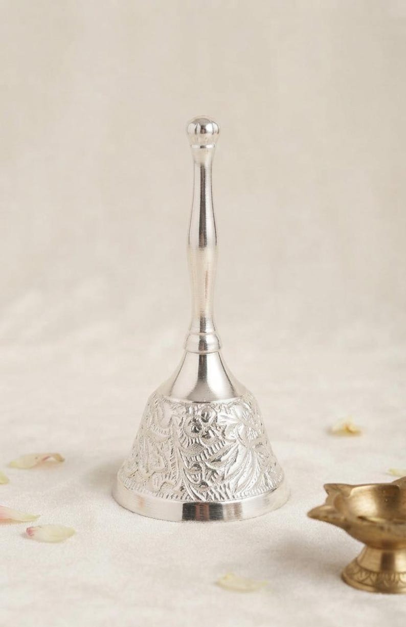 May include: A silver-colored metal hand bell with a long, slender handle and a flared base. The bell features an intricate, embossed floral pattern covering the entire exterior surface of the flared section, ending in a smooth, rounded top knob.