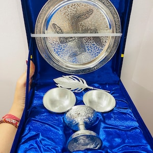 May include: A silver-toned metal set featuring a circular plate with intricate embossed patterns and a central swastika symbol, two small bowls, a feather-shaped spoon, and a stemmed cup, all presented inside a deep blue velvet-lined box.