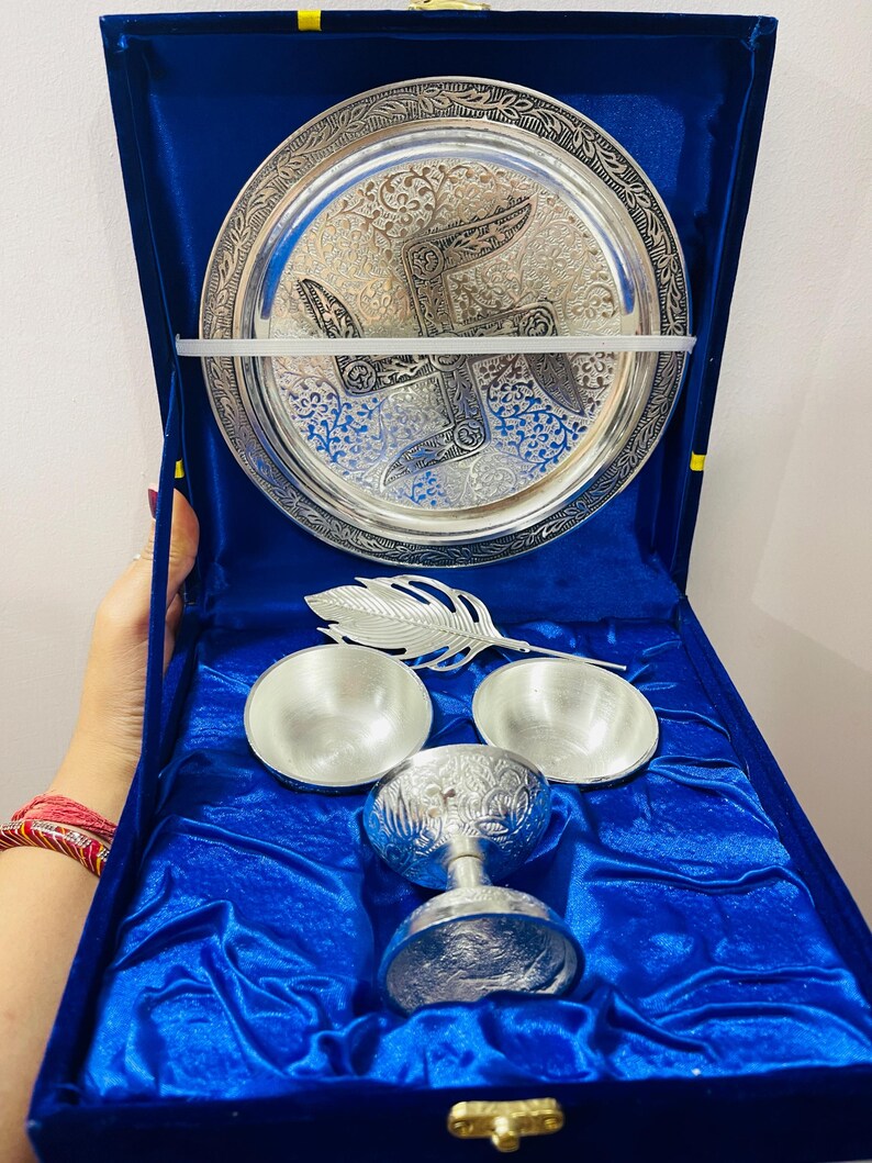 May include: A silver-toned metal set featuring a circular plate with intricate embossed patterns and a central swastika symbol, two small bowls, a feather-shaped spoon, and a stemmed cup, all presented inside a deep blue velvet-lined box.