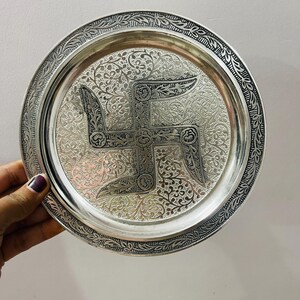 May include: A round silver-toned metal plate featuring an intricate embossed swastika symbol at the center. The surface is decorated with detailed floral patterns and a textured border with a repeating leaf design along the outer rim.
