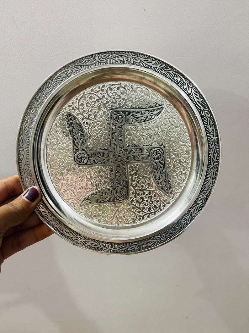 May include: A round silver-toned metal plate featuring an intricate embossed swastika symbol at the center. The surface is decorated with detailed floral patterns and a textured border with a repeating leaf design along the outer rim.