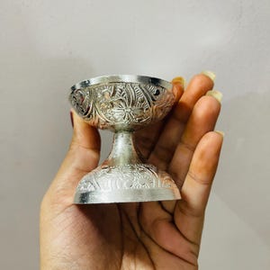 May include: A small, silver-colored metal bowl with a pedestal base, featuring intricate embossed floral and leaf patterns covering the exterior of both the bowl and the base. The object has a polished, reflective finish and a wide, circular rim.