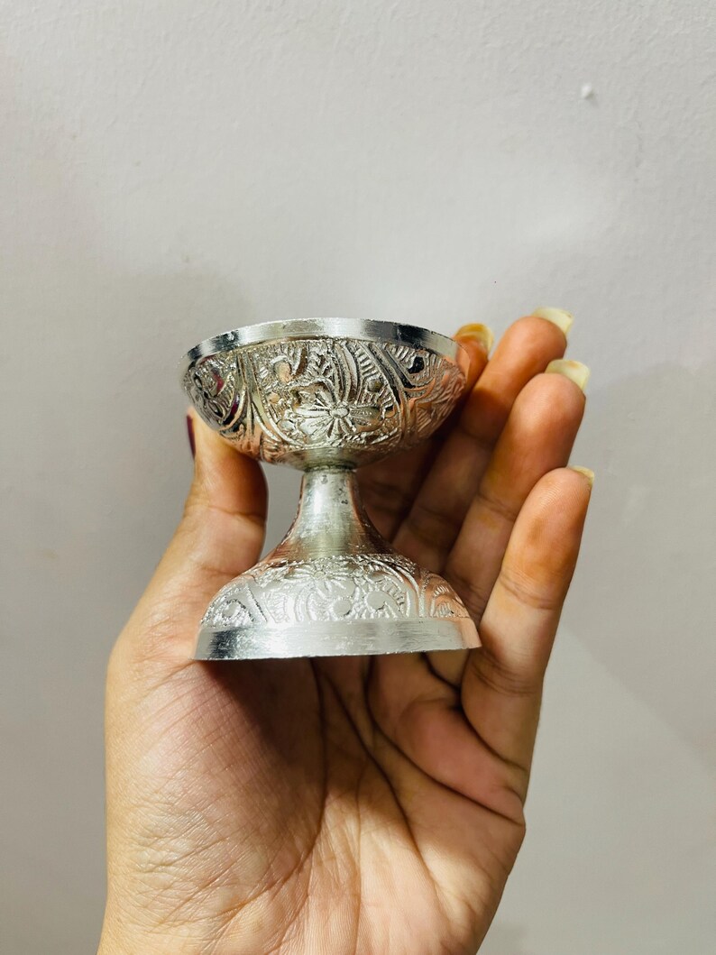 May include: A small, silver-colored metal bowl with a pedestal base, featuring intricate embossed floral and leaf patterns covering the exterior of both the bowl and the base. The object has a polished, reflective finish and a wide, circular rim.