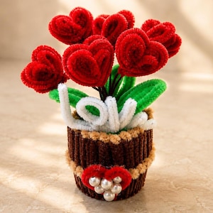 Pode incluir: Um arranjo decorativo feito de limpadores de cachimbo felpudos com várias flores vermelhas em forma de coração e folhas verdes. A palavra LOVE é formada por limpadores brancos na base, e o vaso marrom é decorado com um coração e pérolas.