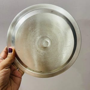 May include: A circular, metallic silver-toned lid with a brushed finish. The lid features a raised outer rim and a slightly recessed center section with a textured, circular pattern radiating from the middle point of the flat surface.