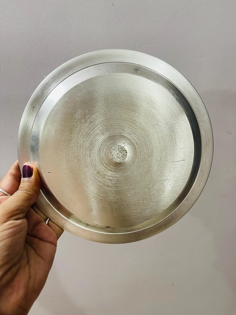 May include: A circular, metallic silver-toned lid with a brushed finish. The lid features a raised outer rim and a slightly recessed center section with a textured, circular pattern radiating from the middle point of the flat surface.