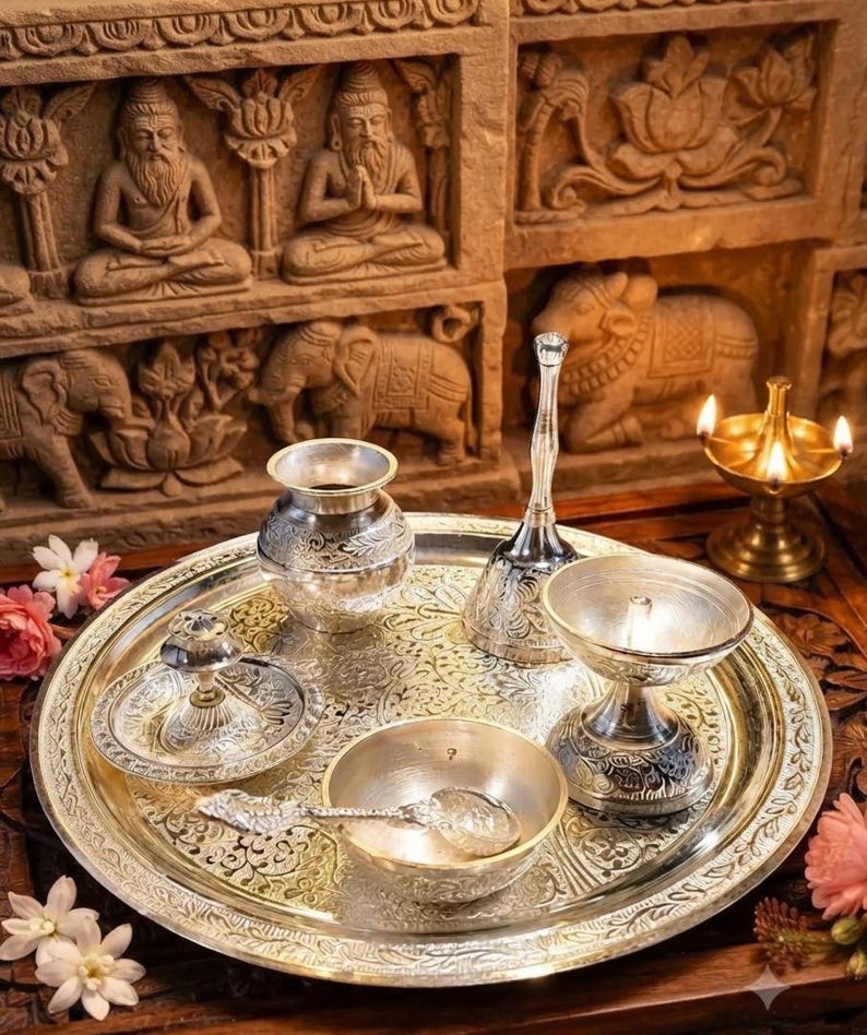 May include: A silver-toned metal puja thali set featuring a large round tray with intricate floral engravings, a small kalash, a bell with a long handle, a diya lamp, a small bowl with a spoon, and a tiny incense holder, all arranged together.