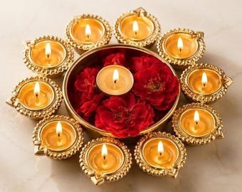 Puede incluir: Una disposición circular de diez lámparas diya de metal dorado, cada una con una vela de té encendida. Un cuenco central dorado está lleno de flores rojas vibrantes y una vela encendida, creando una cálida exhibición de luz y color.