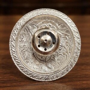 May include: A circular silver-toned metal incense burner featuring an intricate embossed floral pattern across its flat surface. The center of the disc contains a raised, rounded dome with five small circular holes for holding incense sticks.