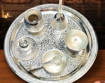 May include: A silver-toned metal puja thali set featuring an ornate floral engraved tray. The set includes a small bell, a lidded container, a shallow bowl with a matching spoon, and a small cup, all displaying intricate embossed patterns.