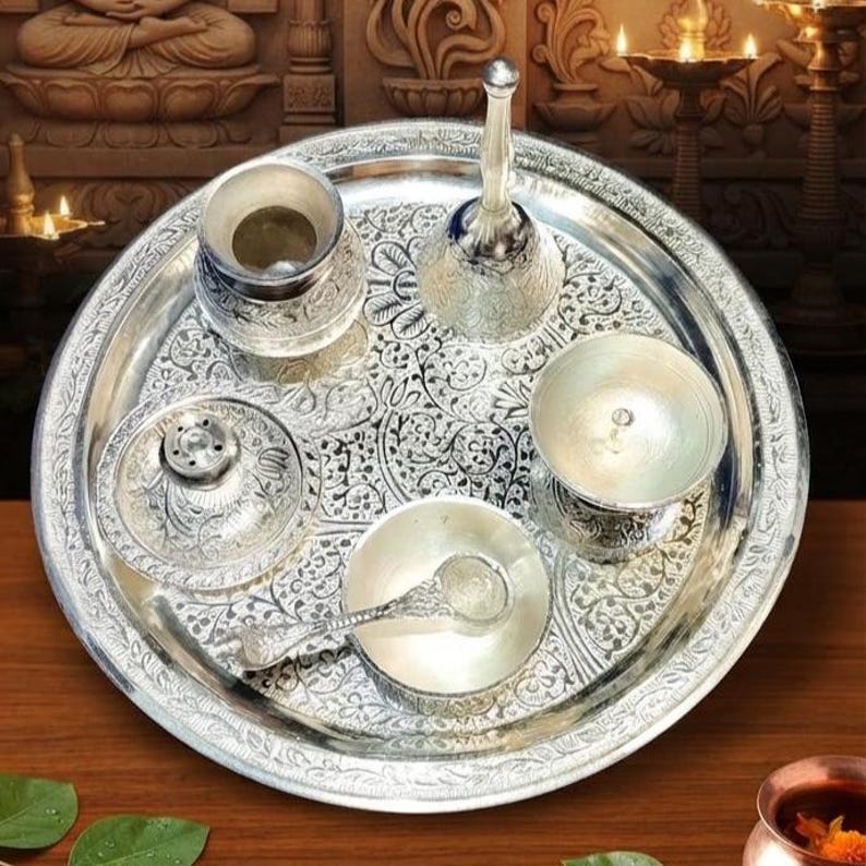 May include: A silver-toned metal puja thali set featuring an ornate floral engraved tray. The set includes a small bell, a lidded container, a shallow bowl with a matching spoon, and a small cup, all displaying intricate embossed patterns.