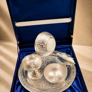 May include: A silver-toned metal puja set featuring a circular tray with intricate floral engravings, a diya lamp, a small bowl, and a leaf-shaped spoon, all presented inside a square box lined with deep blue satin fabric and a gold latch.