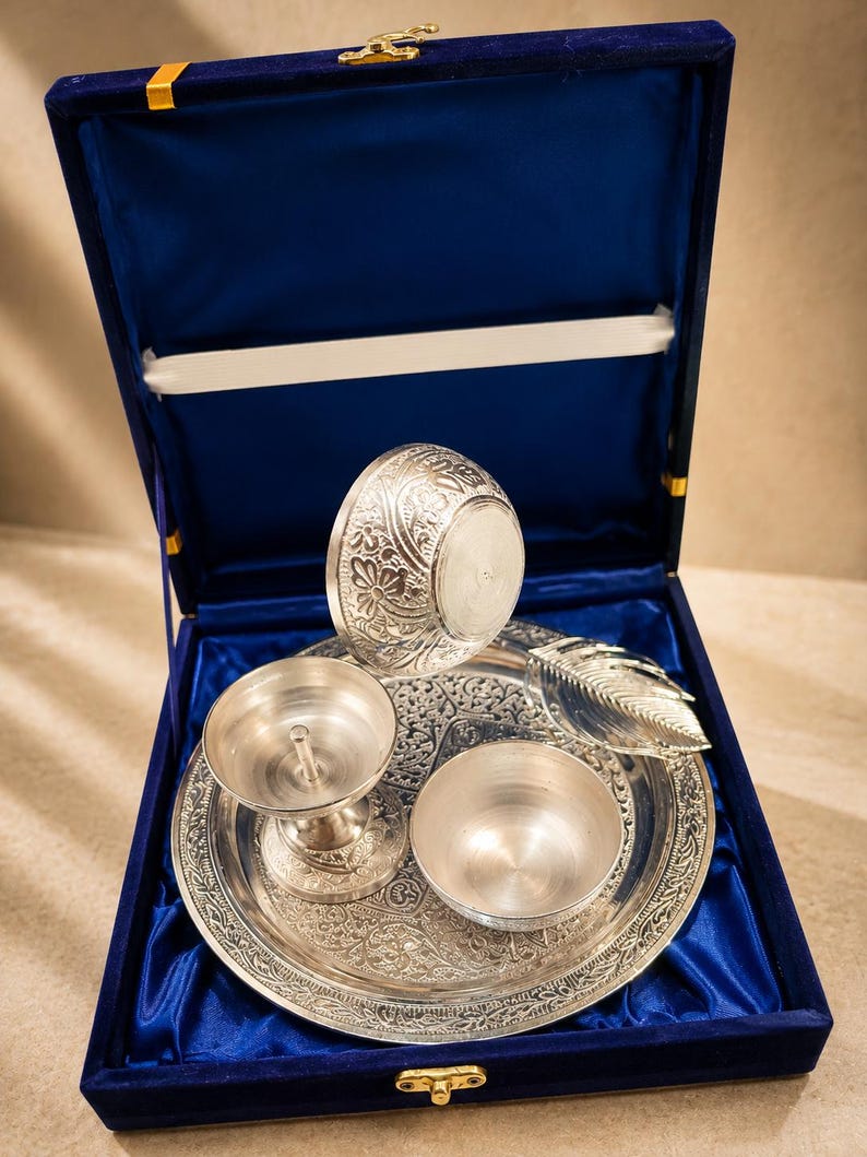 May include: A silver-toned metal puja set featuring a circular tray with intricate floral engravings, a diya lamp, a small bowl, and a leaf-shaped spoon, all presented inside a square box lined with deep blue satin fabric and a gold latch.