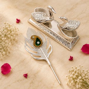 May include: A silver-toned metal set featuring a rectangular base with two swan-shaped containers and a separate feather-shaped object. The feather has a central green paisley-shaped accent with small clear stones and a long, slender handle.